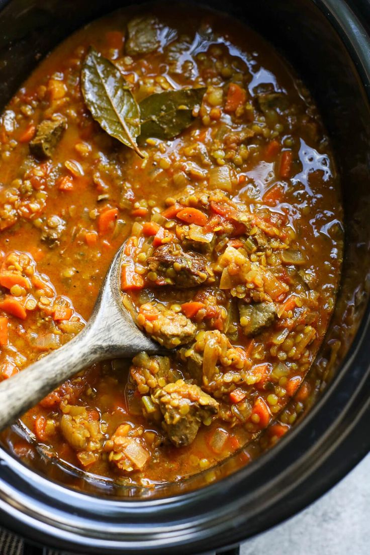 a spoon in a slow cooker filled with lentils and meats, ready to be cooked