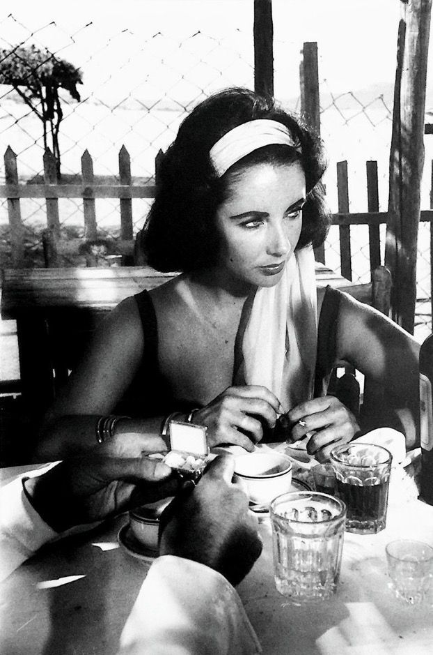 a black and white photo of a woman sitting at an outdoor table with food on it