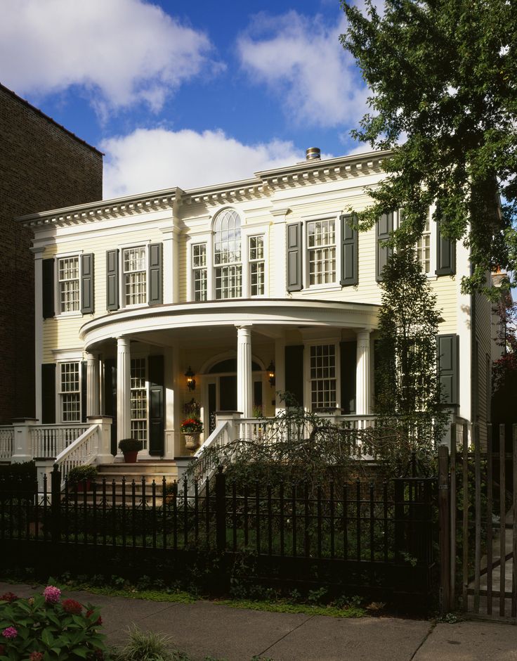 a large white house sitting next to a lush green tree on top of a sidewalk