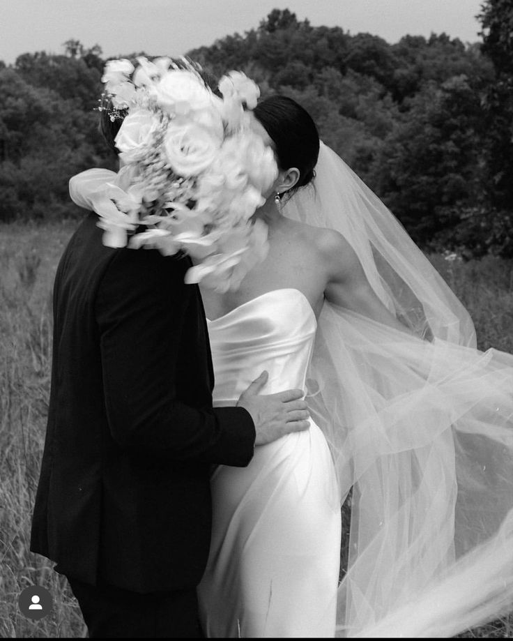 a bride and groom kissing in a field with flowers on their head, veil blowing in the wind