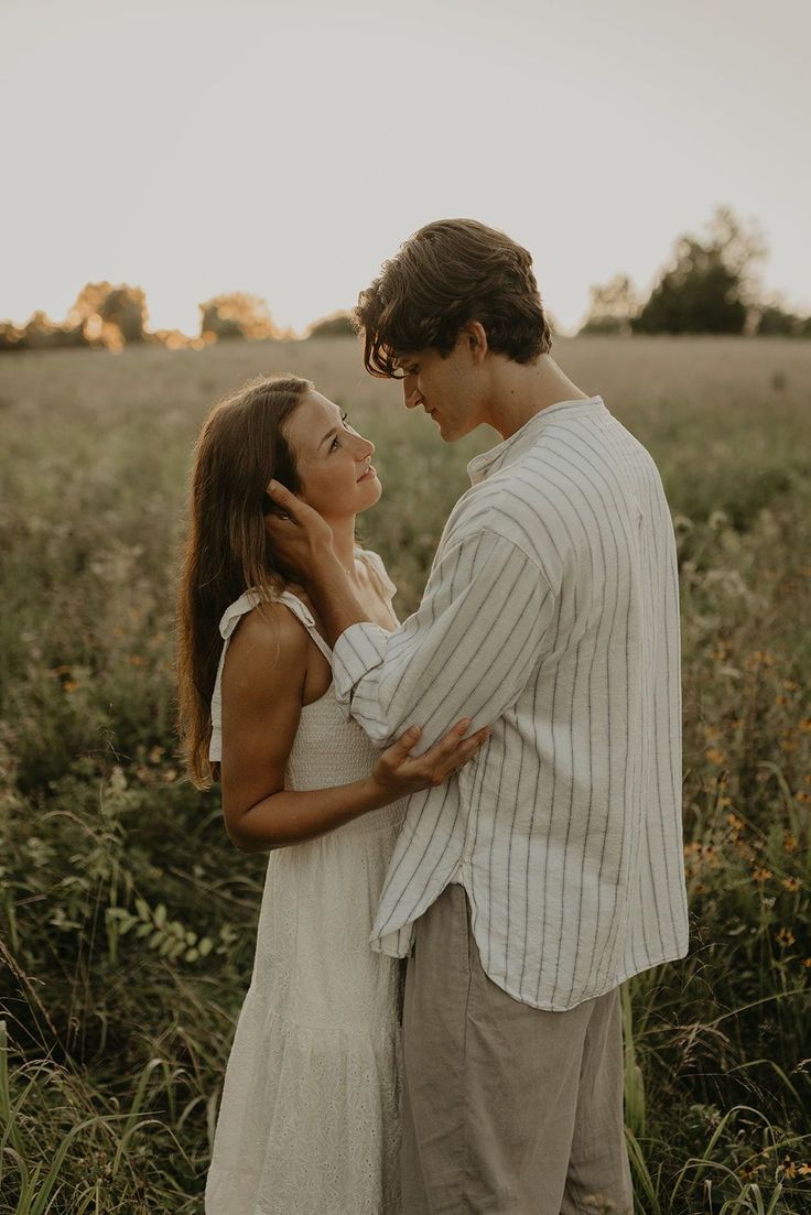 a man and woman standing together in a field with tall grass, looking into each other's eyes