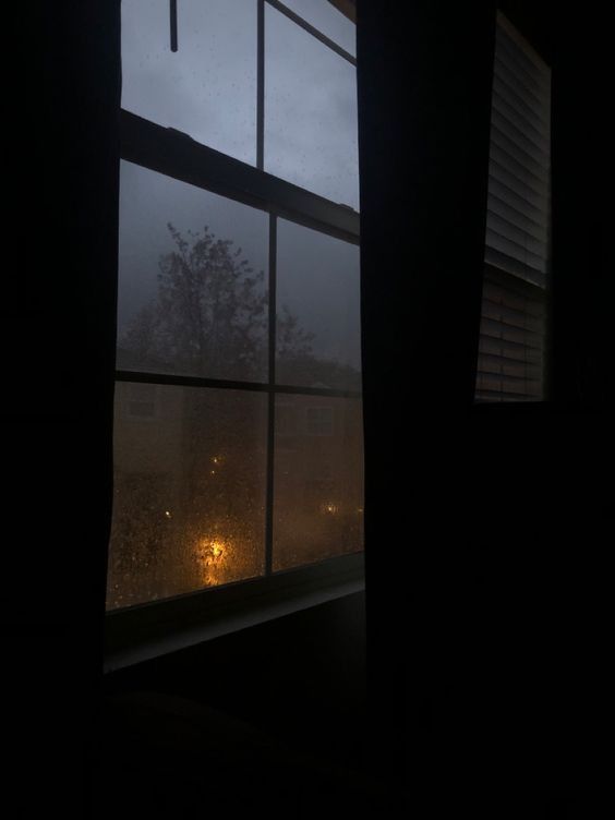 a dark room with an open window and rain coming down on the street lights outside