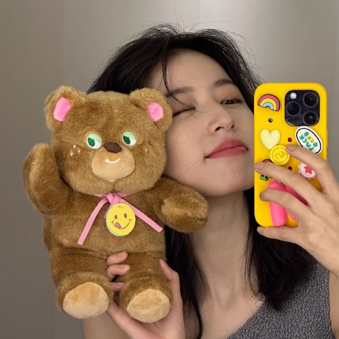 a woman is holding a teddy bear and taking a selfie with her phone