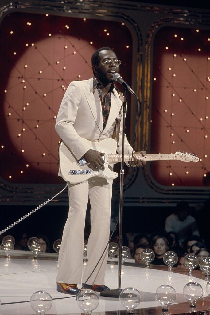 a man in a white suit singing into a microphone and holding a guitar on stage