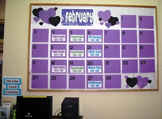 a purple and black calendar with hearts on the wall next to a computer monitor, keyboard and other office supplies