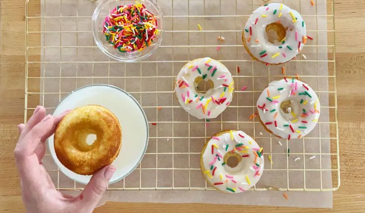 a person holding a donut in front of several doughnuts on a cooling rack