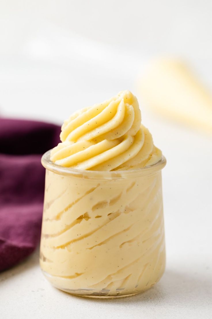 a small glass jar filled with yellow frosting next to a purple cloth on a white surface