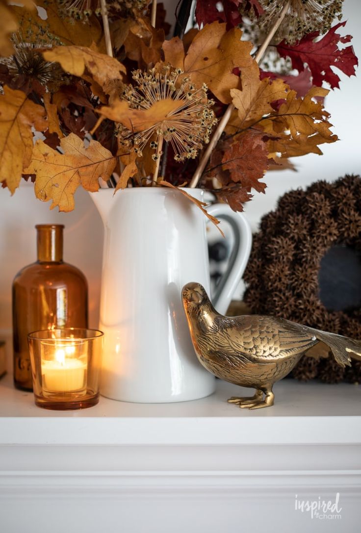 a white vase filled with leaves and a bird figurine on top of a mantle