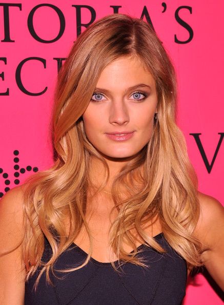 a woman with long blonde hair and blue eyes posing in front of a pink wall