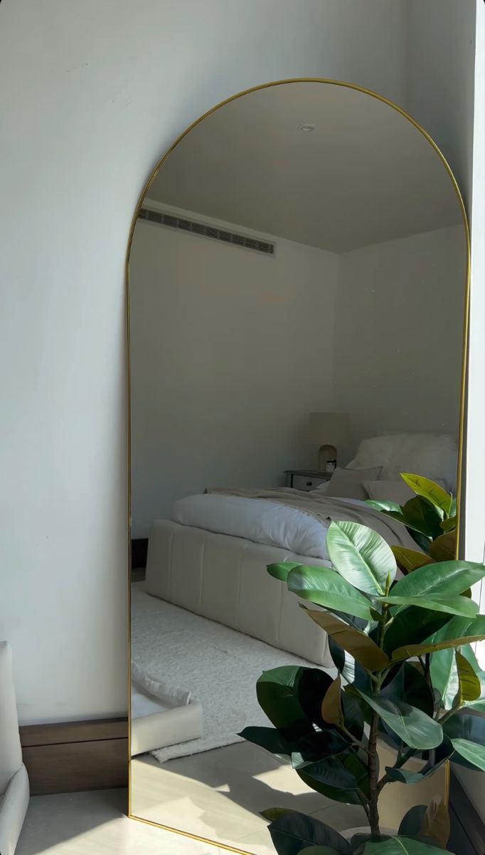 a mirror with a plant in it next to a white chair and bed on the other side