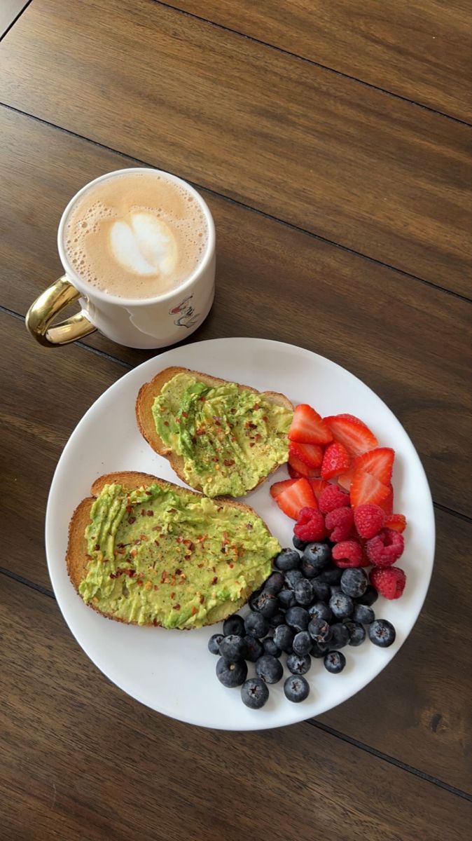 an avocado toast with strawberries and blueberries on a plate next to a cup of cappuccino