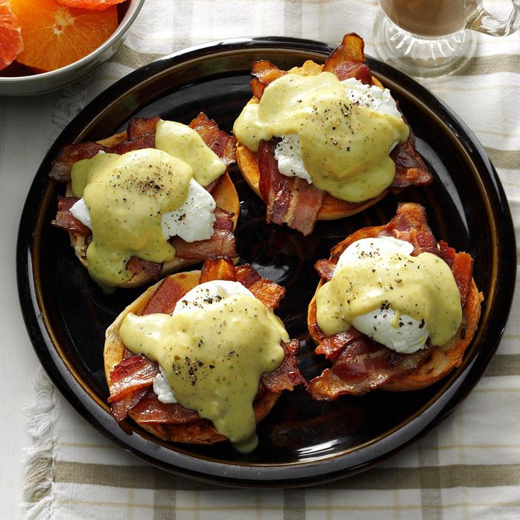 eggs benedict with bacon and hollandaise sauce on a black plate next to an orange slice