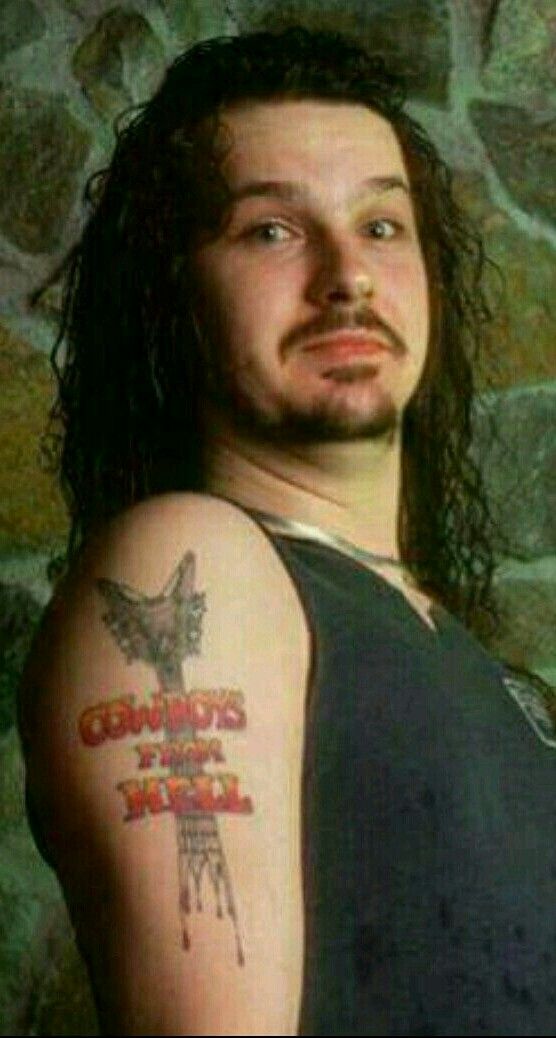 a man with long hair and a goat tattoo on his arm, standing in front of a stone wall