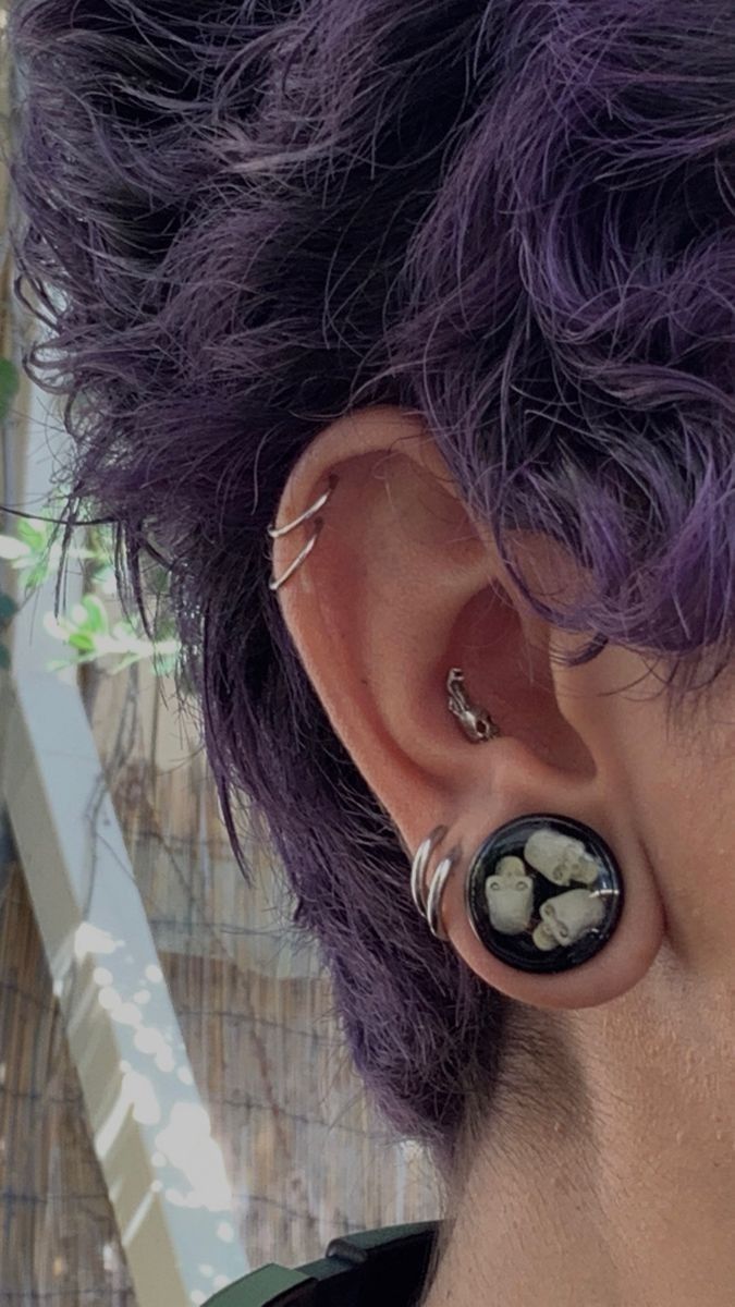 a woman with purple hair wearing ear rings and piercings on her left ear is looking away from the camera