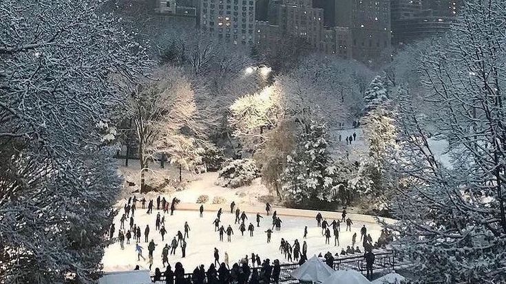 people skating on an ice rink in the middle of a snowy park with tall buildings