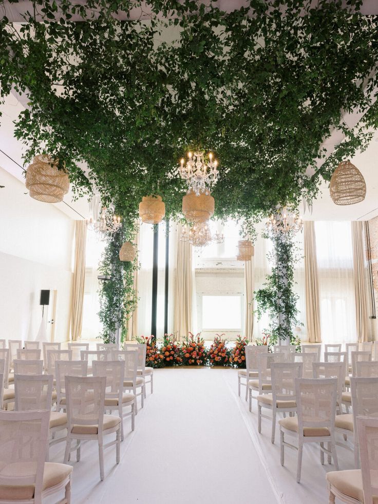 an indoor wedding venue with white chairs and greenery on the ceiling, chandeliers hanging from the ceiling