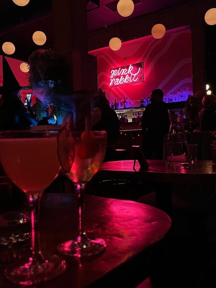 two wine glasses sitting on top of a table in front of a neon lit bar