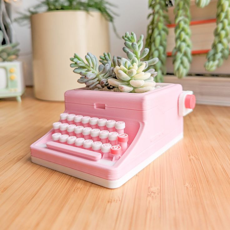 a pink toy typewriter sitting on top of a wooden table next to succulents