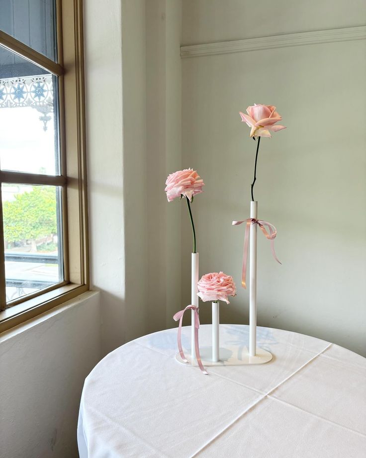 two white vases with pink flowers sitting on a round table in front of a window