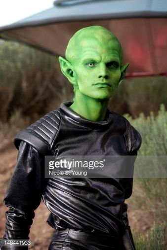 a green man in black leather outfit standing on dirt road next to grass and bushes