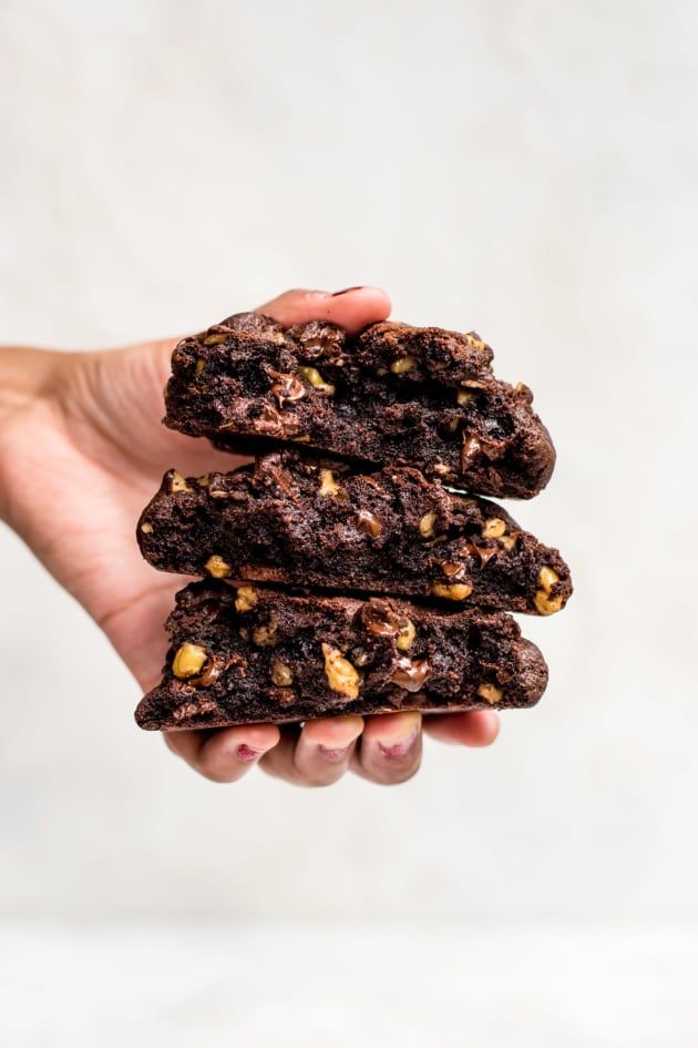 a hand holding three chocolate cookies with nuts on top in front of a white background