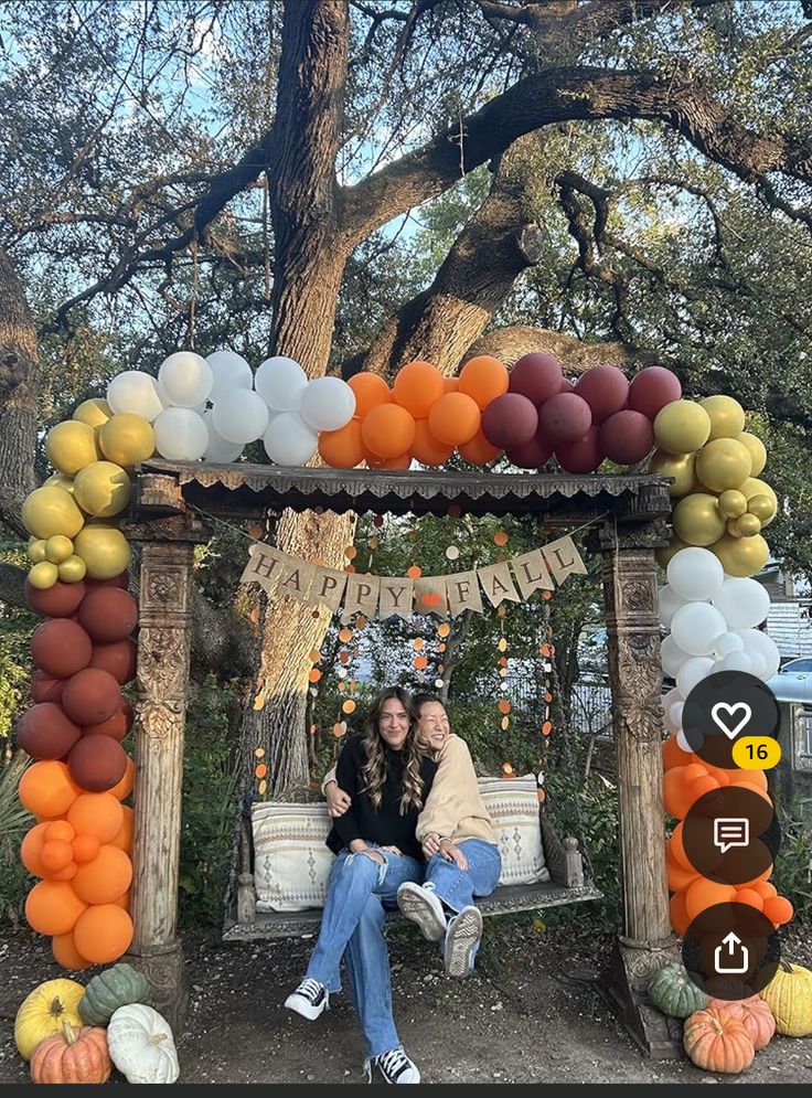 two women sitting on a bench in front of an arch made out of balloons and pumpkins