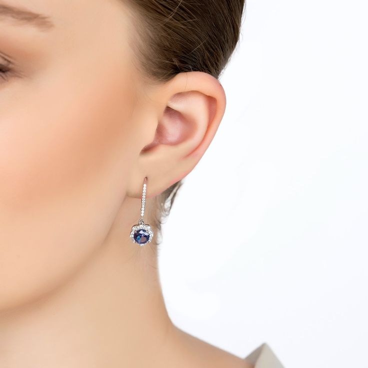 These earrings feature a beautiful floral-inspired design. Each earring has a round, lab-grown tanzanite stone surrounded by sparkling cubic zirconia forming a delicate flower. The secure latch back fastener, adorned with additional zirconia, adds extra sparkle. Tanzanite is the birthstone for December. It symbolizes transformation, intuition, and spiritual growth. The Matilda Drop Earrings make a thoughtful gift for birthdays, especially December birthdays, anniversaries, or any special occasio Elegant Tanzanite Earrings With Brilliant Cut, Tanzanite Earrings With Prong Setting, Tanzanite Round Earrings For Wedding, Round Tanzanite Earrings For Weddings, Tanzanite Round Wedding Earrings, Elegant Diamond Birthstone Earrings, Round Tanzanite Wedding Earrings, Elegant White Gold Diamond Earrings With Birthstone, Elegant Sapphire Earrings With Sparkling Stones