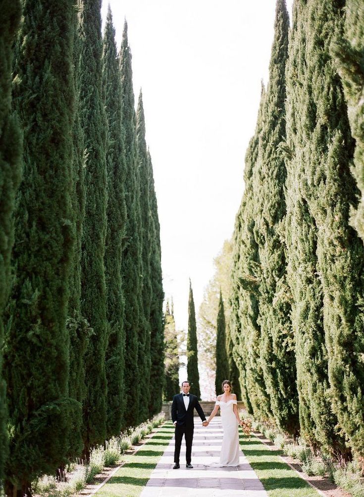 a bride and groom walking down a path lined with tall trees in front of them