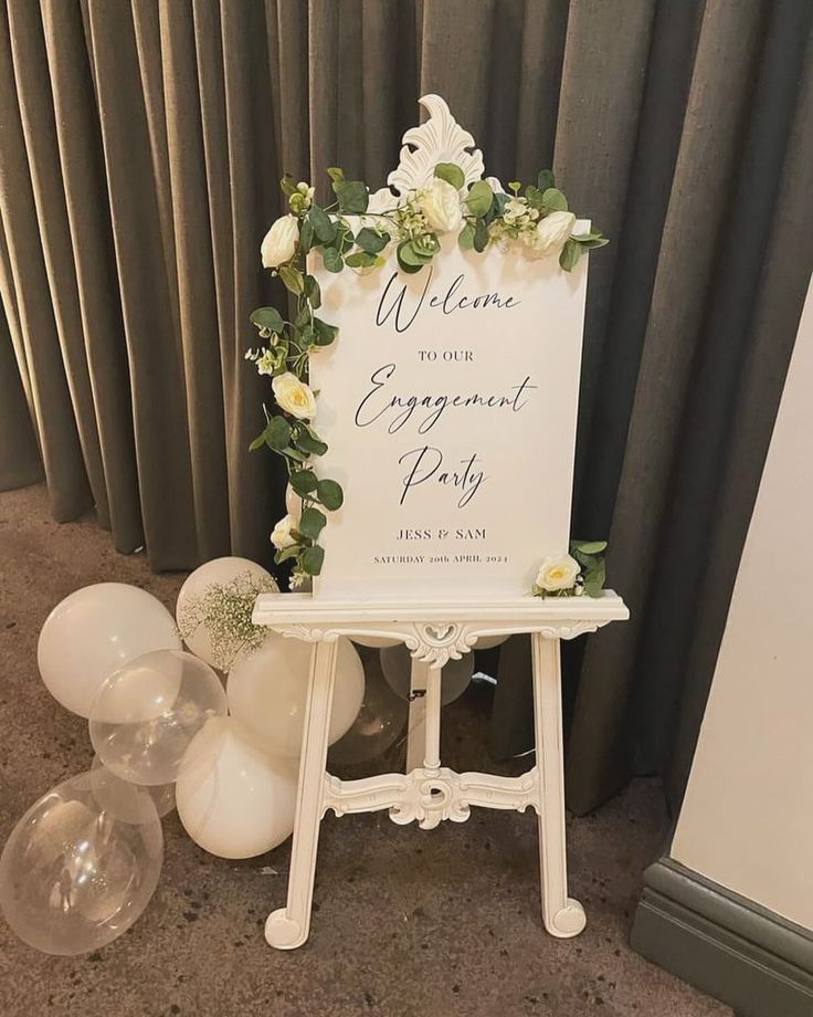 a welcome sign with flowers and greenery sits on an easel next to balloons