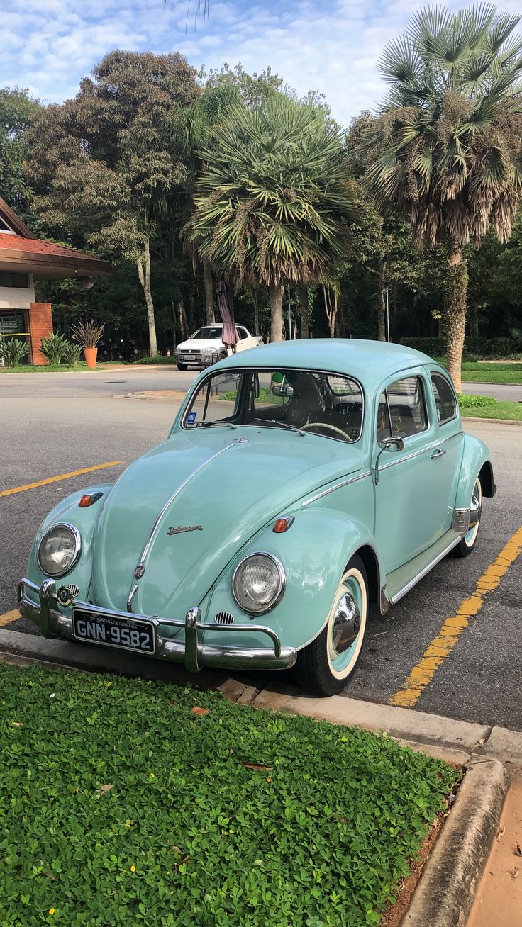 an old blue car parked in a parking lot next to some trees and grass on the side of the road