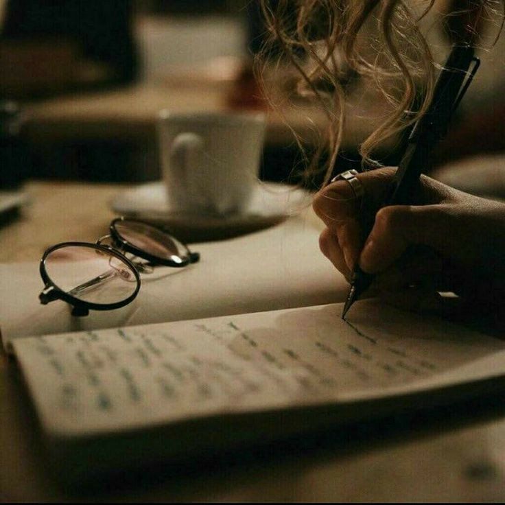 a person writing on a piece of paper next to a coffee cup and eyeglasses