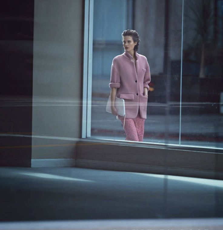 a woman in pink is standing by a window with her hand on her hip and looking out the window