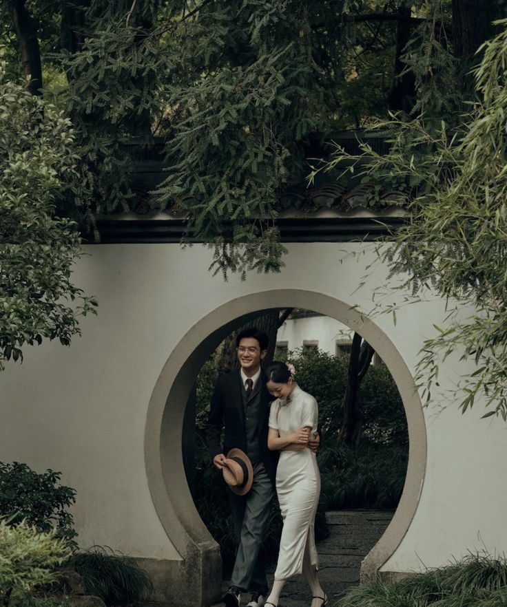 a man and woman are standing in front of a circular structure with trees around them