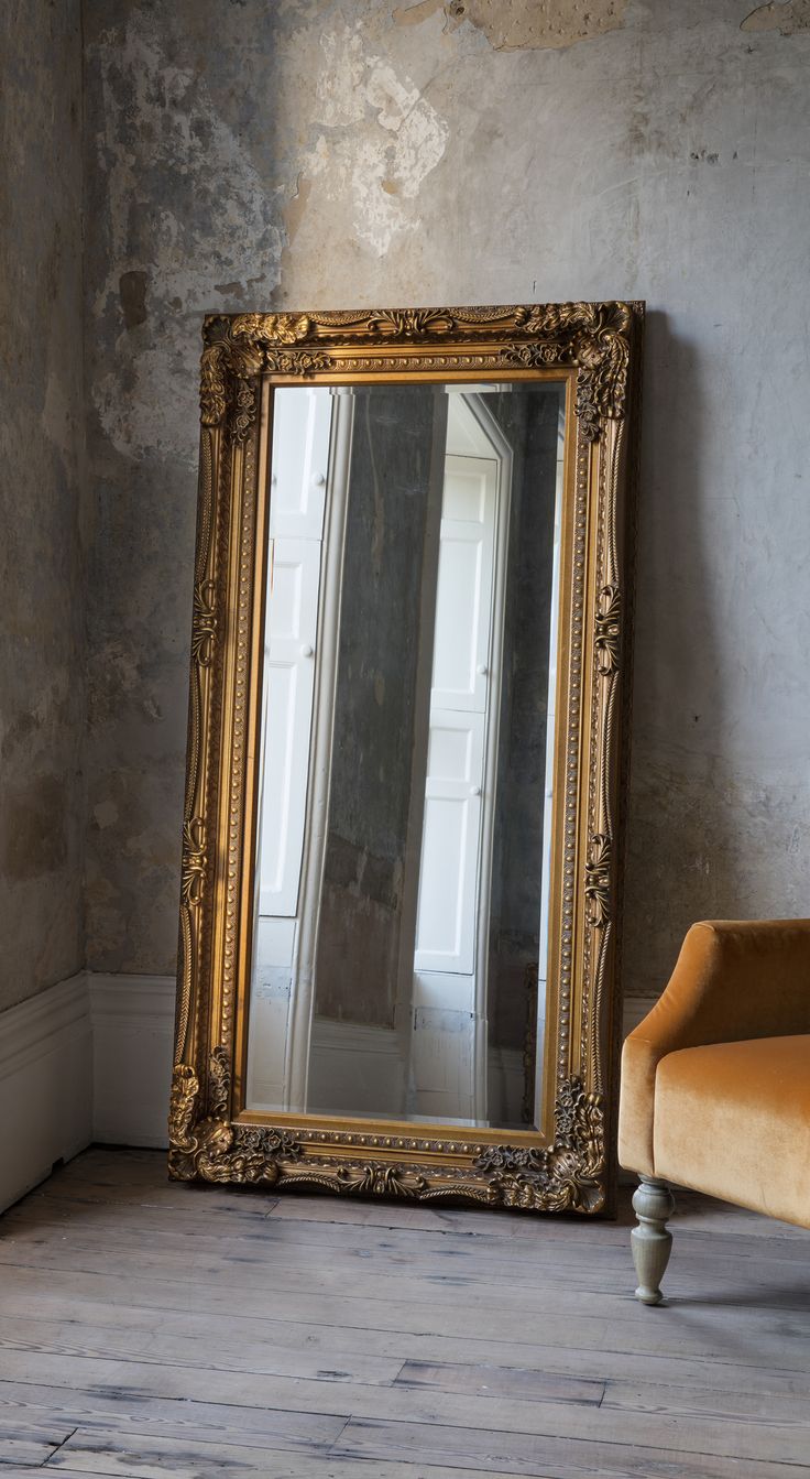 a large gold framed mirror sitting on top of a wooden floor next to a chair