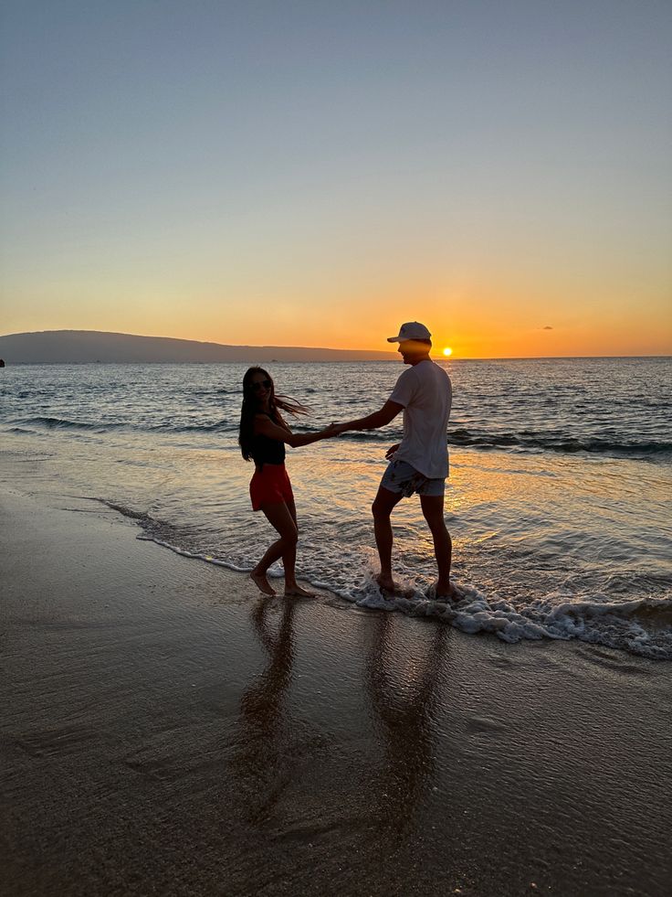 a man and woman holding hands on the beach at sunset with waves lapping in front of them