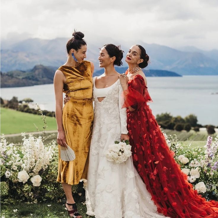 three women standing next to each other in front of some flowers and water with mountains in the background