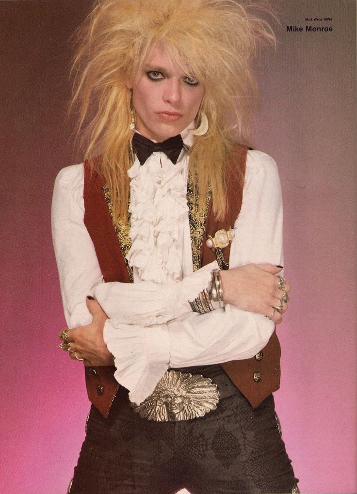 a woman with long blonde hair standing in front of a pink background wearing a white shirt and brown vest