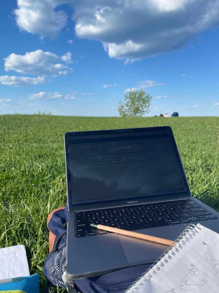 an open laptop computer sitting on top of a grass covered field next to a notebook