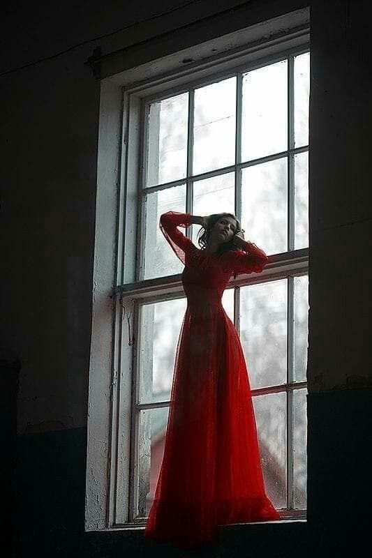 a woman in a long red dress standing by a window with her hands on her head