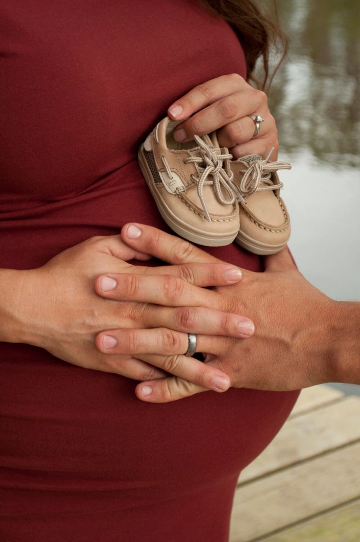 a pregnant woman holding her baby's shoes while standing next to a body of water