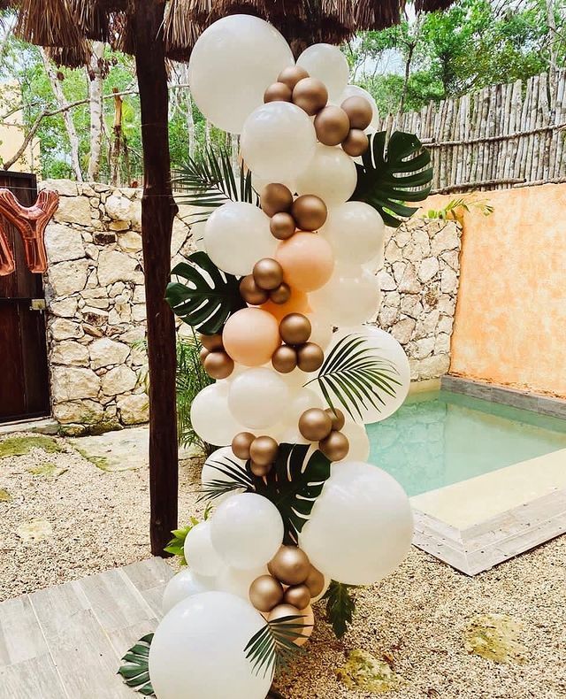 some white and gold balloons are hanging from a palm tree near a small swimming pool