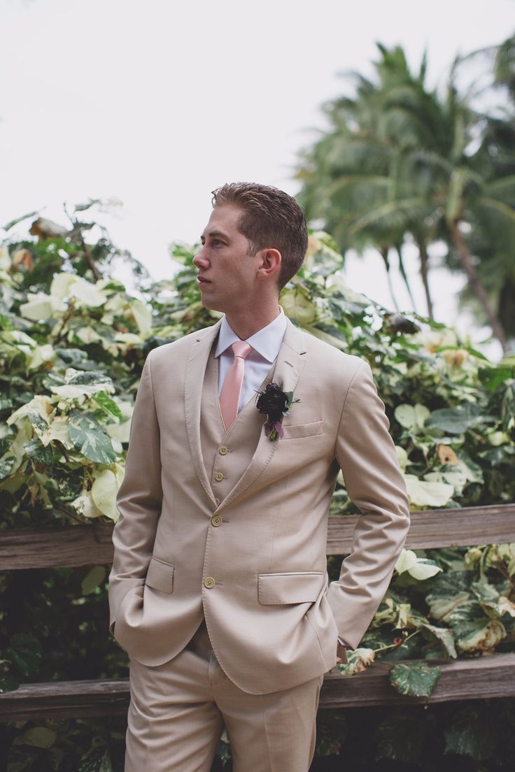 a man wearing a tan suit and pink tie standing next to a wooden bench in front of palm trees