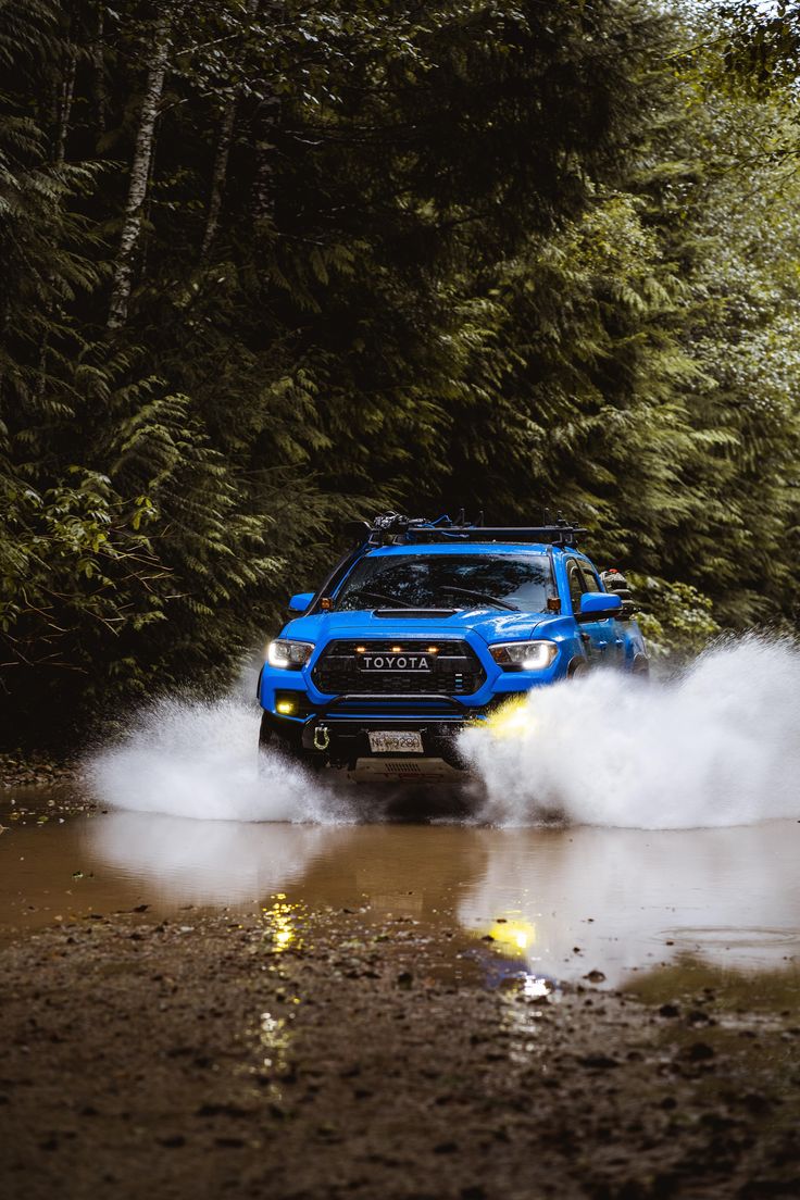 a blue truck is driving through the mud in front of some trees and water with its lights on