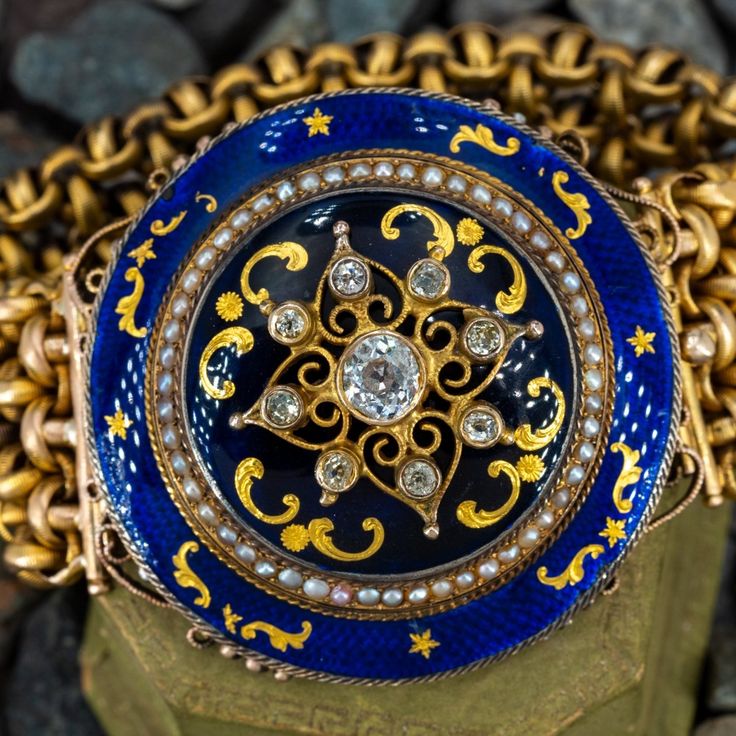 This antique bracelet features a Victorian era 10K yellow gold diamond and pearl domed setting (converted from a brooch) accented with blue enamel and gold accents. The bracelet is accented with nine (9) old European/old mine cut diamonds and fifty-two (52) seed pearls. The seed pearls measure an average of 1.7mm. The 14K yellow gold fancy mesh link bracelet measures 25mm wide and 6.1mm thick. The bracelet measure an overall length of 7 inches and is finished with a hidden box clasp. The bracelet has natural patina and light enamel loss. It fits up to a 6.5 inch wrist. Luxury Jeweled Yellow Gold Jewelry, Antique Blue Jewelry With Brooch, Antique Jeweled Jewelry, Antique Enamel Bracelet Collectible, Formal Jeweled Yellow Gold Jewelry, Formal Medallion Brooch Jewelry, Formal Yellow Gold Jeweled Jewelry, Blue Intricate Design Jewelry Bracelet, Collectible Enamel Bracelet Jewelry