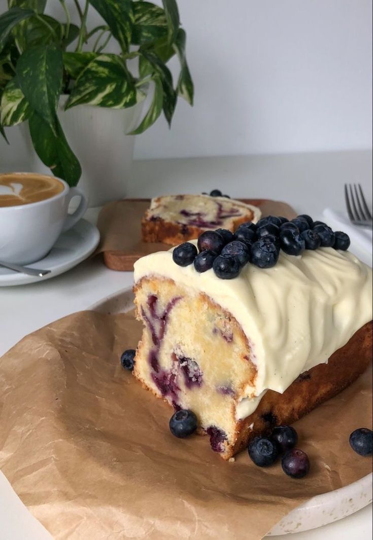 a piece of cake with blueberries and cream frosting sitting on a plate next to a cup of coffee