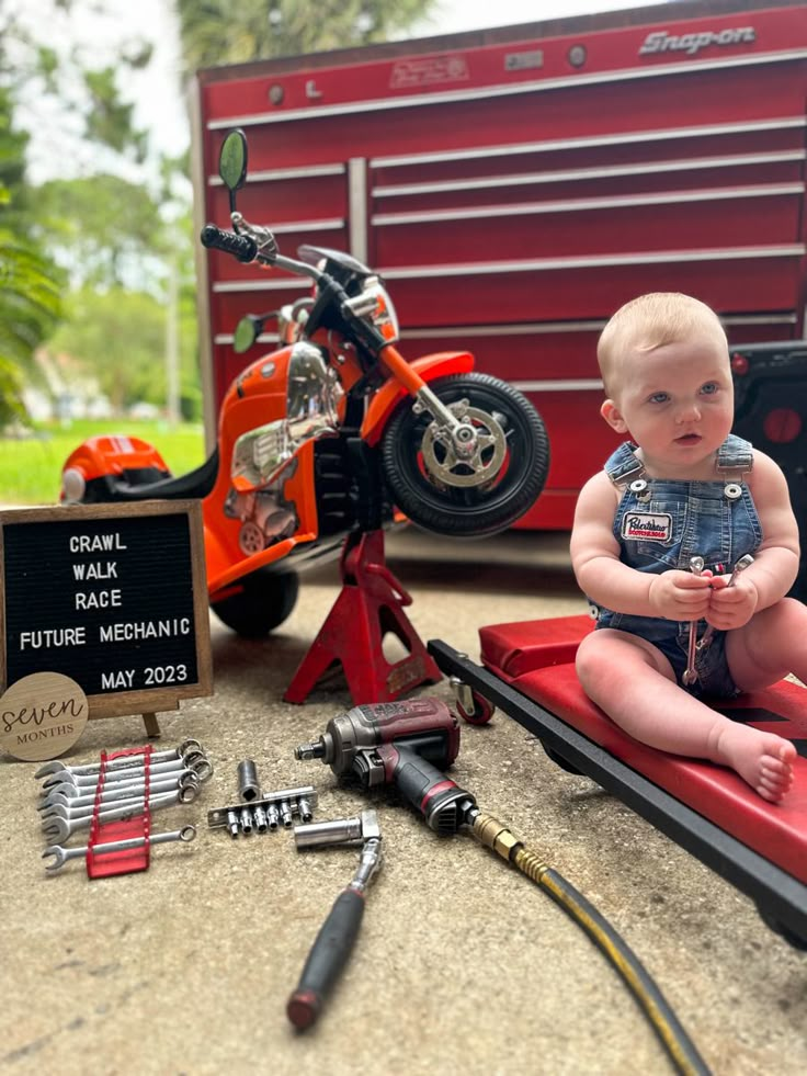 a baby sitting on top of a red box next to tools and a motorbike