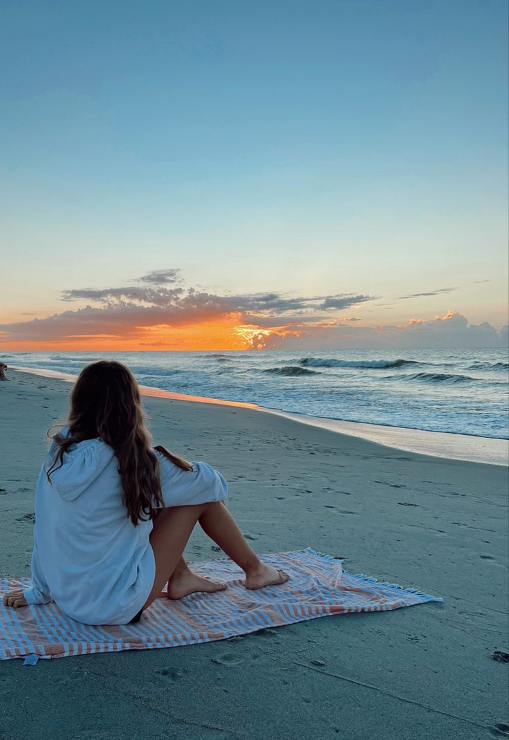 a woman sitting on top of a towel at the beach watching the sun go down