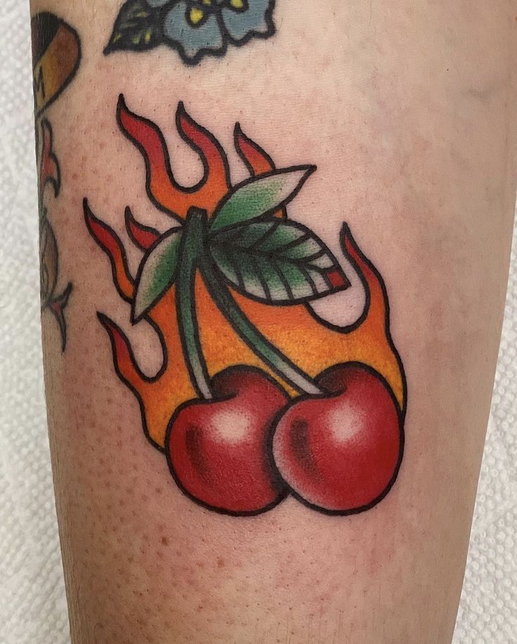 a tattoo with cherries and flames on the thigh, as well as flowers in the background