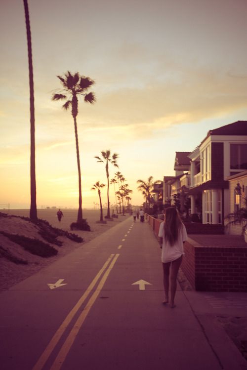 a woman walking down the street in front of some palm trees at sunset or dawn