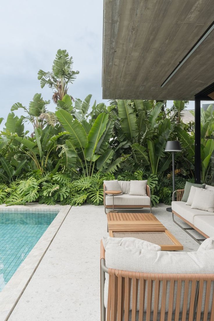 an outdoor living area with couches and tables next to a swimming pool surrounded by greenery